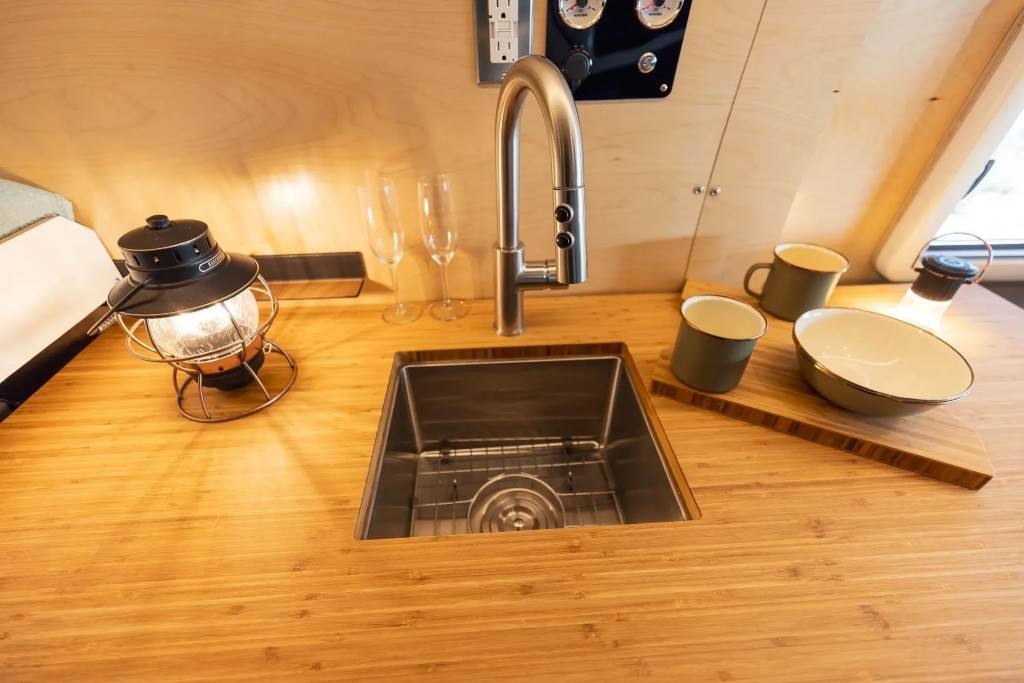 A close-up of a DIY camper van sink setup, showing a fresh water tank, gray water tank, and PVC tubing connected to a drop-in sink
